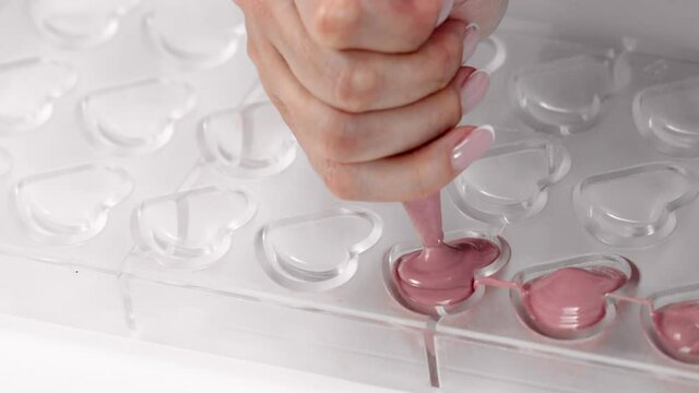 Chocolate production, candymaking concept - confectioner filling heart shape candy mold with melted ruby premium chocolate, making hand-crafted candies. Confectionery small family factory, slow motion
