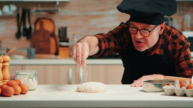 Elderly chef with bonete making dough baking cookies. Retired senior baker with apron, kitchen uniform sprinkling, sifting, spreading rew ingredients with hand baking homemade pizza and bread.