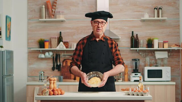 Old man wearing bonete while explaining food preparation recipe looking at camera. Retired blogger chef influencer using internet technology communicating on social media with digital equipment
