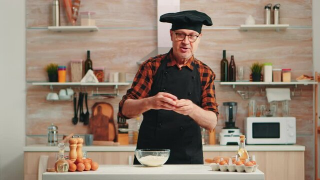 Experienced baker man craking eggs for baking wearing apron enjoying hobby. Retired elderly chef with bonete mixing by hand, kneading in glass bowl pastry ingredients preparing homemade cake