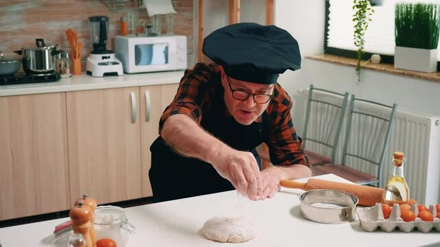 Retired baker man sieving flour on chopping board making dough at home. Elderly senior chef with bonete and uniform sprinkling, sifting, spreading ingredients by hand, baking homemade pizza and bread