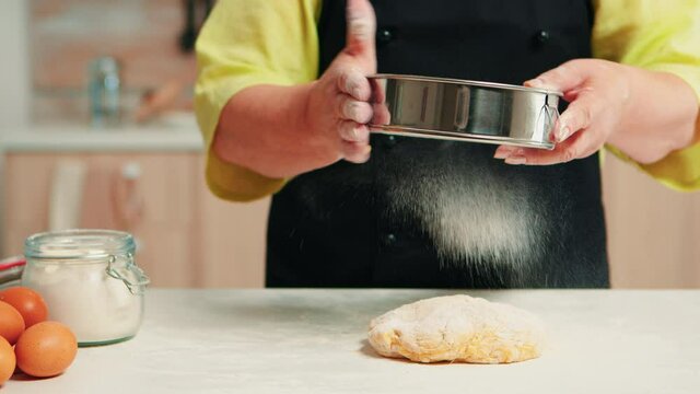 Senior woman with black apron sifting flour on dough using metallic sieve Happy elderly chef with bonete preparing raw ingredients to baking traditional bread sprinkling, sieving in the kitchen.