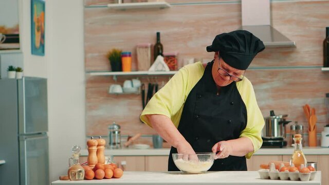 Mature woman baker mixing by hand cracked eggs with flour in home kitchen following traditional recipe. Retired elderly chef with bonete kneading in glass bowl pastry ingredients baking homemade cake