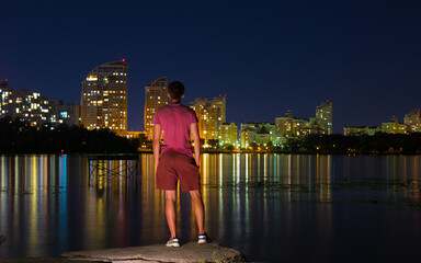Fototapeta na wymiar A guy stands on the banks of the Dnieper river against the background of a bright night embankment