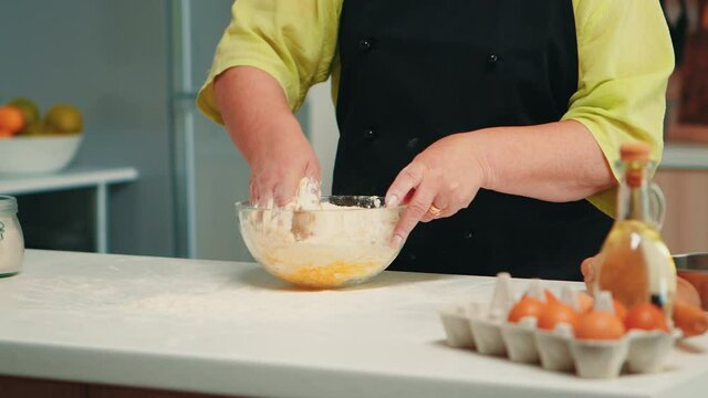 Chef senior woman mixing by hand bread ingredients in home kitchen in glass bowl. Retired elderly baker with bonete kneading cracked fresh eggs with wheat flour baking homemade cake and bread