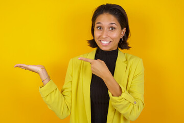 Young hispanic businesswoman wearing casual turtleneck sweater and jacket pointing and showing empty copy space on open hand palm for text