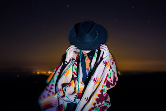 Woman in Blanket Holds Her Hat Against Soft Starry Sky and Night Cityscape