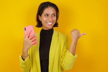 Young hispanic businesswoman wearing casual turtleneck sweater and jacket using and texting with smartphone  pointing and showing with thumb up to the side with happy face smiling
