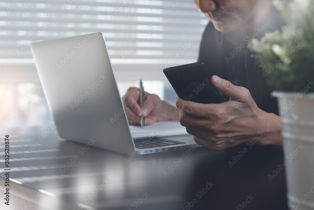 Poster man using mobile phone and writing on notepad while working on laptop computer in office