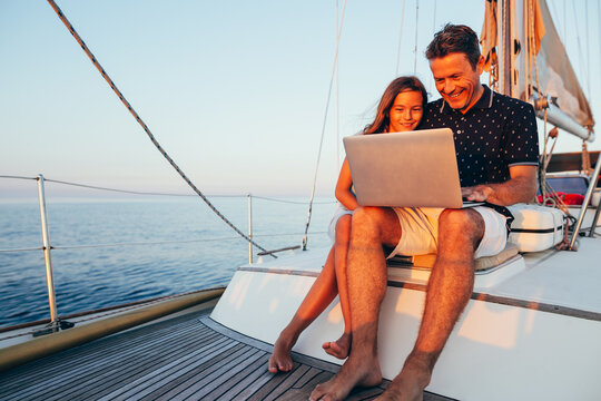 Father and daughter using laptop on a sailboat at sunset.