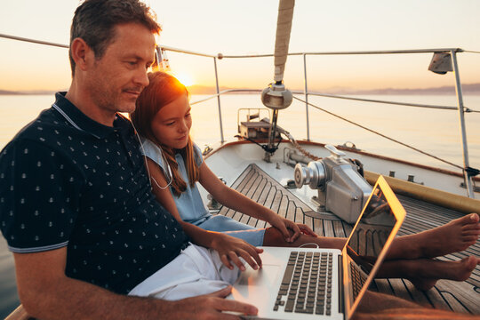 Father and daughter using laptop on a sailboat at sunset.