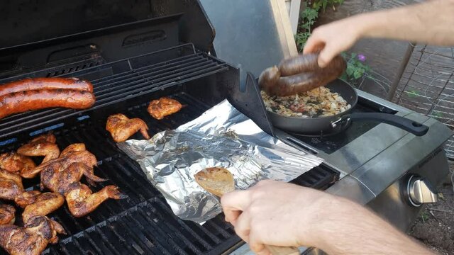Barbecuing food like chicken wings and sausage while transferring and placing browned barley or blood sausage into previously sated chopped onion in skillet on side burner.