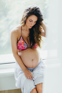 Beautiful woman with pregnant belly sits at the window in bikini and sarong skirt