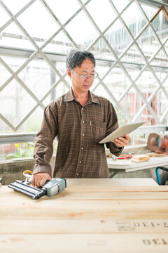 Male Wood Worker Looking at Tablet Computer and Holding Nail Gun in Bright Wood Shop