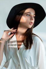 Fashion look. Romantic casual style. Beautiful young woman in white shirt black wide-brimmed hat posing in sun light shadow isolated on neutral. Season trend. Female elegance.