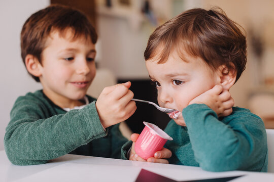 Young boy helping his little brother to eat a strawberry yoghurt.