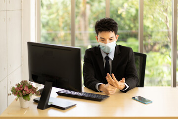 A young asian businessman in suit wearing face mask for protective covid-19 sits at his desk in the office give himself a hand massage after working on a computer all day.