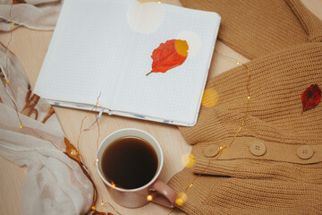 A Cup of coffee, an open diary on empty pages lie on the table, on which lies a warm knitted sweater of brown color, a white patterned scarf and an led garland that is twisted in a spiral on the table