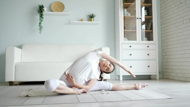 Attractive pregnant lady in white clothes does janu sirsasana position sitting on floor with rug against designer couch in spacious room at home