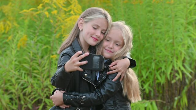 Portrait of Cute Little Girls make selfie photo on smartphone in a beautiful flowering field. Happy little kids using smartphone playing mobile game, browsing social media.