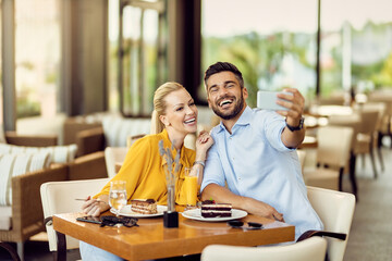 Happy couple having fun while eating dessert and taking selfie in a cafe.
