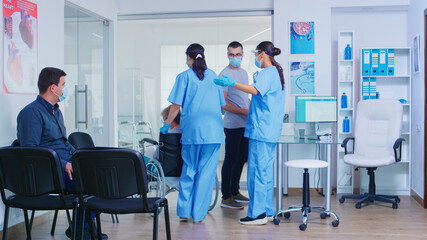 Nurse wearing visor against covid-19 in hospital waiting area talking with patient. Assistant pushing disabled senior woman in wheelchair wearing face mask and sterile gloves.