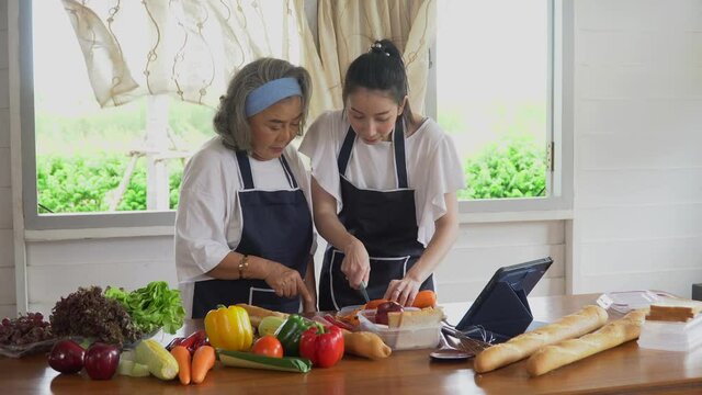 happy asian young daughter and senior mother cooking online  class on tablet together making fresh vegetables food in kitchen at home