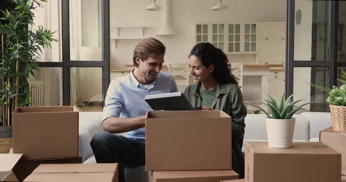 Happy millennial couple sit on couch in living room open unpacking cardboard box with personal belongings after relocation or house renovation. Happy homeowners at moving day at first new home concept