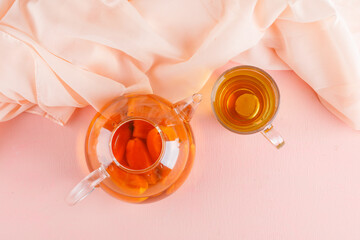 Apricot tea in teapot and glass mug on pink and textile background, top view.