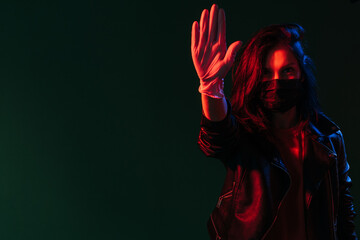Pandemic restriction. Social distancing. Street style woman in black protective face mask glove showing stop gesture in red blue neon light isolated on dark night teal copy space background.