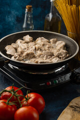 Close-up of preparing pork meat in frying pen on the stove. Dark blue background. Backstage of cooking meal with meat at the professional restaurant kitchen. Vertical format. Food concept.