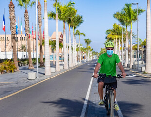 Senior man with medical mask and helmet riding in middle of the deserted street his bicycle in Tenerife, Canary islands. No tourism due to Covid-19 coronavirus. Palm trees and blue sky in background