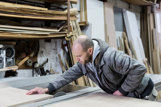 
Young carpenter passing wood board through industrial table saw