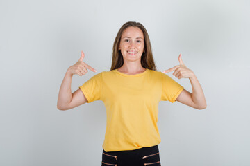 Young woman showing herself with fingers in yellow t-shirt, pants and looking cheery. front view.