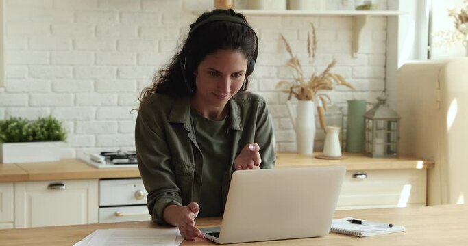 Female work from home standing in kitchen negotiating with business partner remotely wear headset use laptop and video call conference app. E-learning with e-tutor distantly, modern tech usage concept