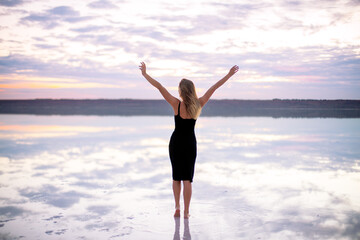 Fototapeta na wymiar beautiful girl in a black dress on a salty pink lake at a vanilla sunset raised her hands