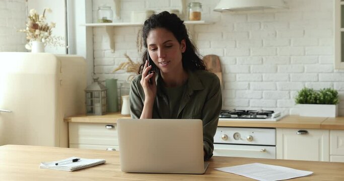 Woman sit at table in kitchen talk on phone to consultant before make order online remotely, businesswoman texting email on laptop chatting with colleague enjoy pleasant conversation working from home