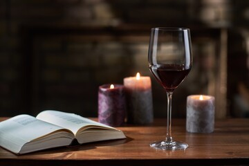 Glass of red wine, open book with candles in the background on wooden table.