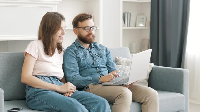 Happy husband and wife relaxing sitting on sofa enjoying using laptop computer, young couple looking at notebook screen browsing internet choosing shopping online resting together in apartment