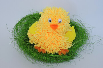 A cute yellow fluffy handmade Easter chicken with orange beak and legs and yellow wings, a chick made of yellow yarn sitting in a green nest made of sisal fibers, white background