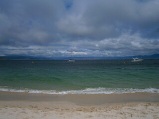 Waves against the sand under a cloudy sky with boats on the ocean