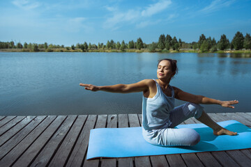 A young woman sits on a blue mat on a wooden dock and makes torso turns with her arms wide apart. Meditation, yoga in nature.