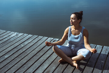 A young woman on a wooden pier sits in a lotus position with her head turned to the side with her eyes closed. Meditation, yoga in nature.