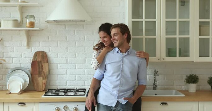 Couple talk spend time in kitchen, wife sit on kitchen counter top hug from behind husband, planning future life together at new modern house. Bank loan for young family, love romantic date concept
