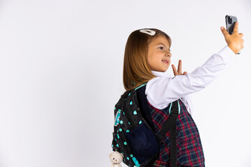 Photo of joyful beautiful schoolgirl taking selfie on cellphone and gesturing peace sign isolated white background