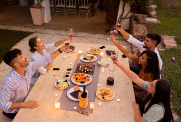 Top view of group of friends toasting with red wine glasses at sunset on a big white granite table in the garden laden with delicacies and lit candles
