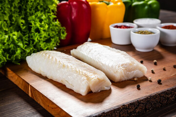 Fresh raw cod with seasonings and vegetables served on cutting board on wooden table
