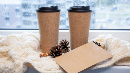 A cup for coffee near the window, a knitted scarf, pine cones, a letter. Autumn and winter background.