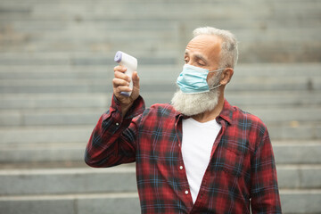 Bearded senior man in face mask holding infrared thermometer a healthy senior man's normal body...