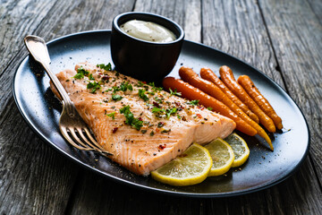 Fried salmon fillet with carrots, lemon and cream sauce served on black plate on wooden table
